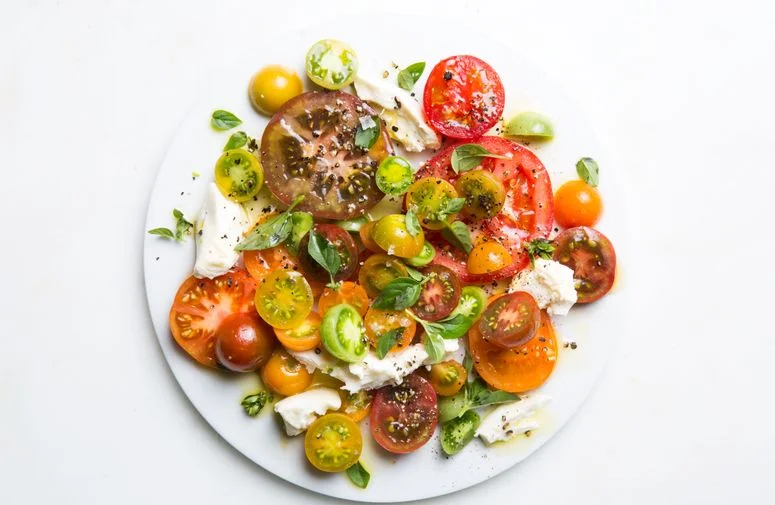 Overhead shot of Caprese salad on a white platter with various types of tomatoes, torn mozzarella, and small basil leaves.