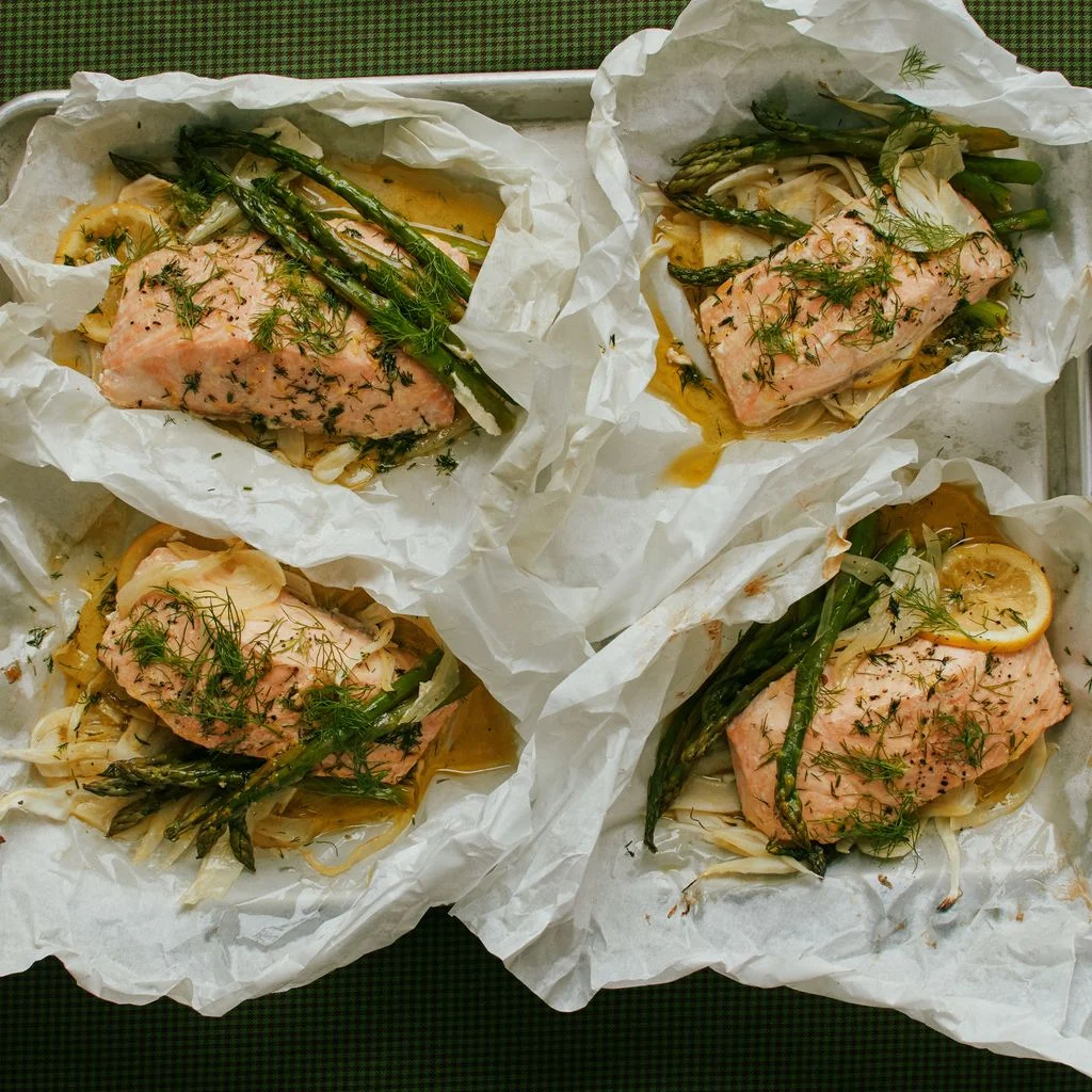 Salmon in papillote on a sheet tray with roasted asparagus fennel lemon slices and topped with fresh dill and black pepper.