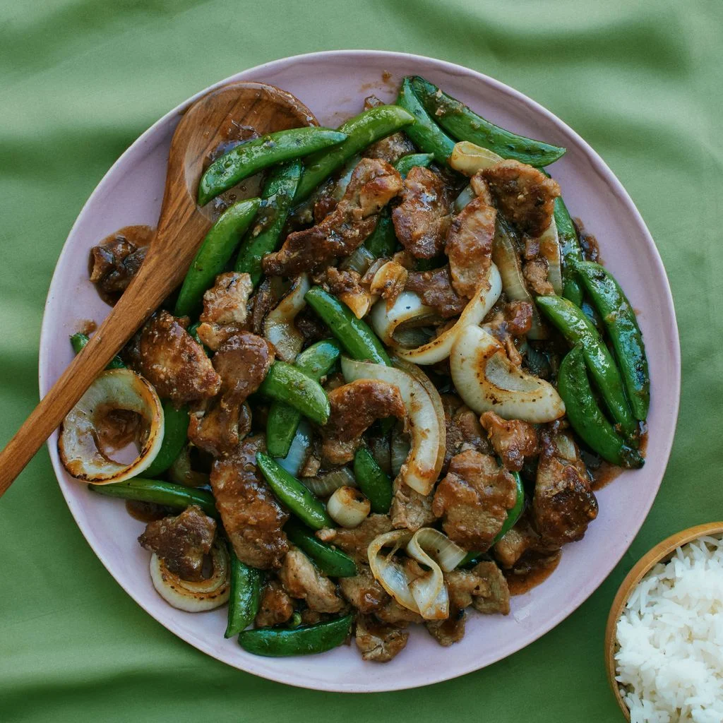 Ginger glazed pork snap peas garlic and onions on a serving platter with bowls of white rice.