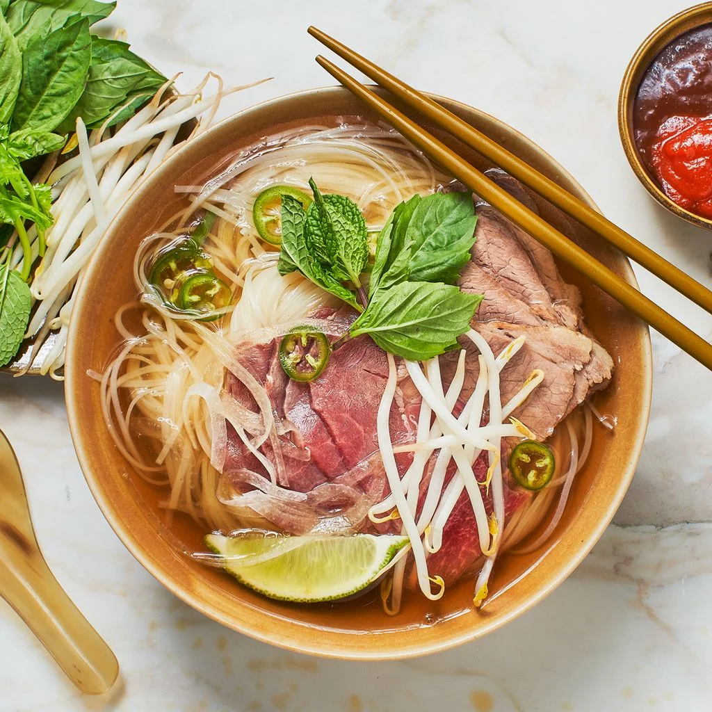 A bowl of beef pho shot from above topped with bean sprouts and Thai basil with condiments to the side.