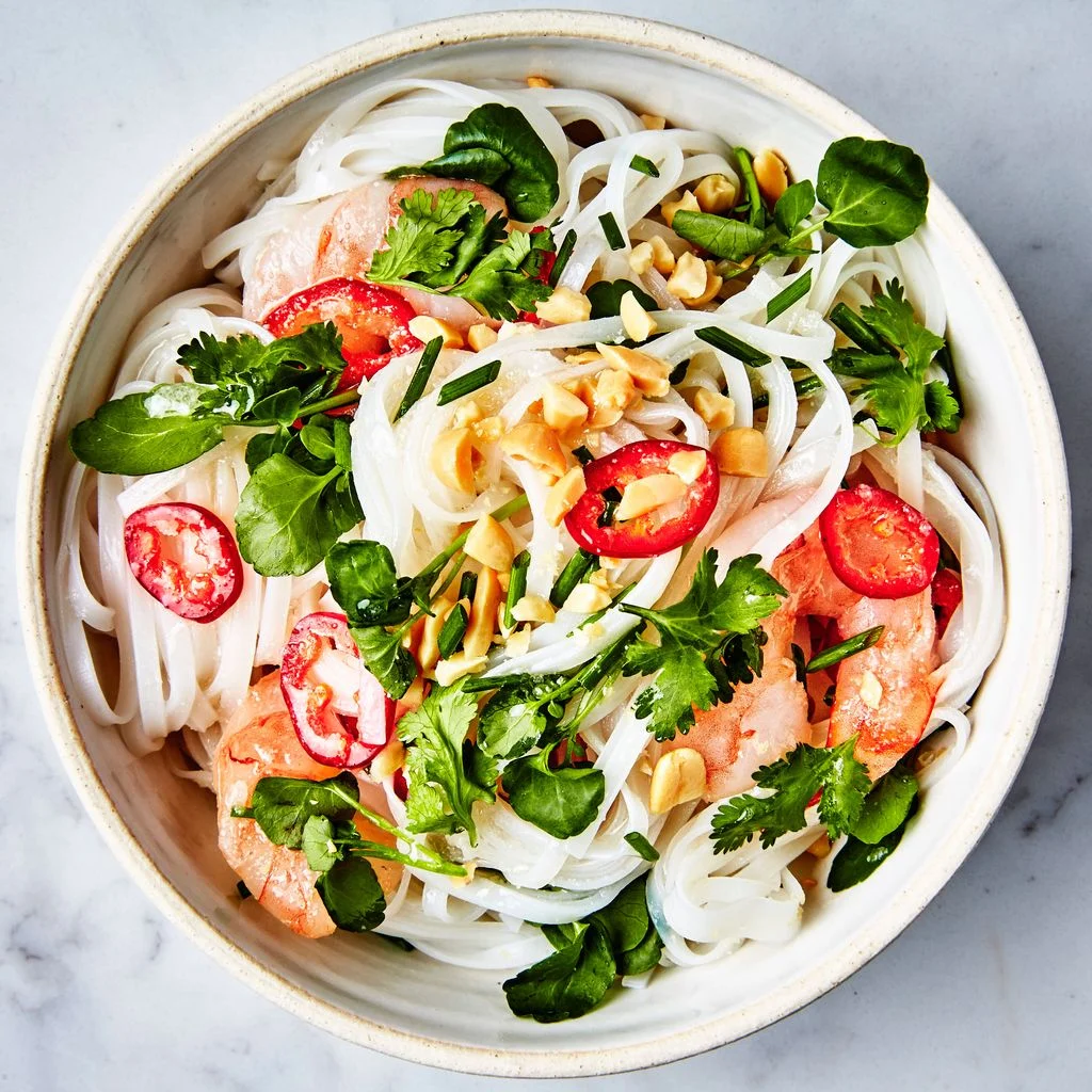 A bowl of rice noodles tossed with shrimp sliced red chiles herbs and chopped peanuts.