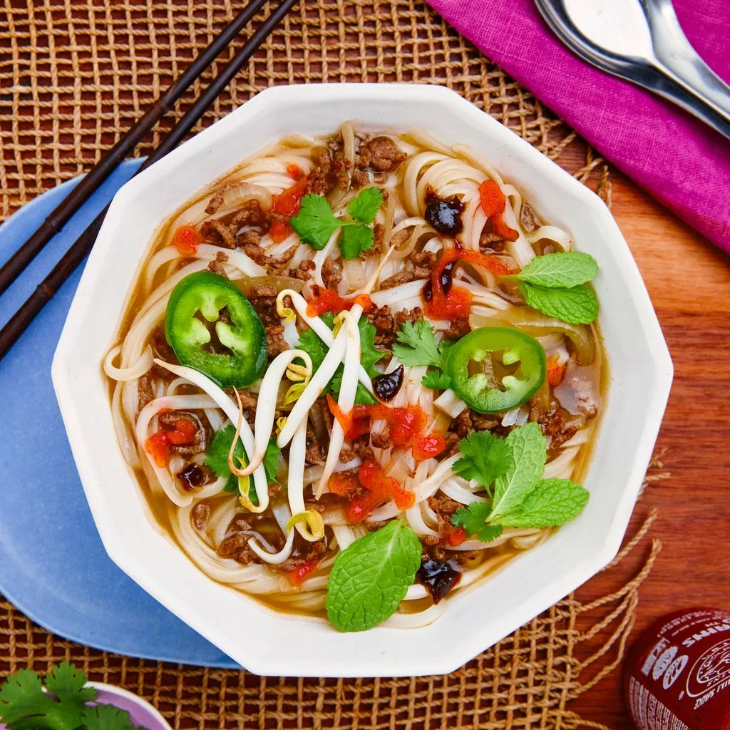 A bowl of vermicelli ground beef sliced jalapeños mint leaves and sriracha in a broth.